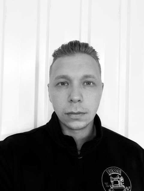 Black and white headshot of a person wearing a black jacket against a white striped background, looking directly at camera