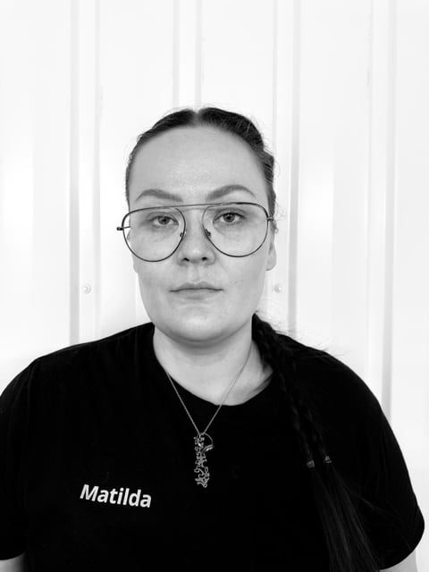 Black and white portrait of a person wearing round glasses and black shirt with name Matilda, against white vertical blinds background